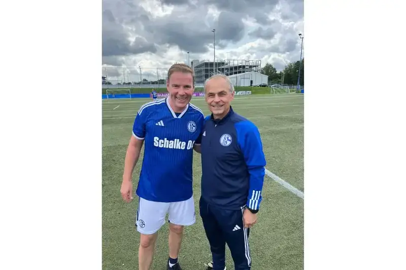Two men smiling for a photo on an outdoor soccer pitch; one wears a blue Schalke 04 jersey and white shorts, while the other wears a dark blue Adidas team tracksuit.