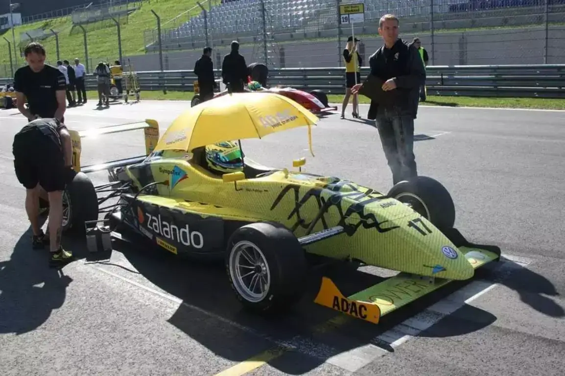 A yellow and black Zalando-sponsored formula race car sits on a sunny track with a driver in the cockpit shaded by a yellow umbrella while crew members perform final checks.