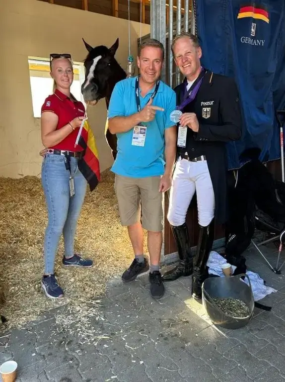 Three people, including a rider in a black competition coat holding a silver medal, pose with a dark horse in a stable adorned with a German flag.