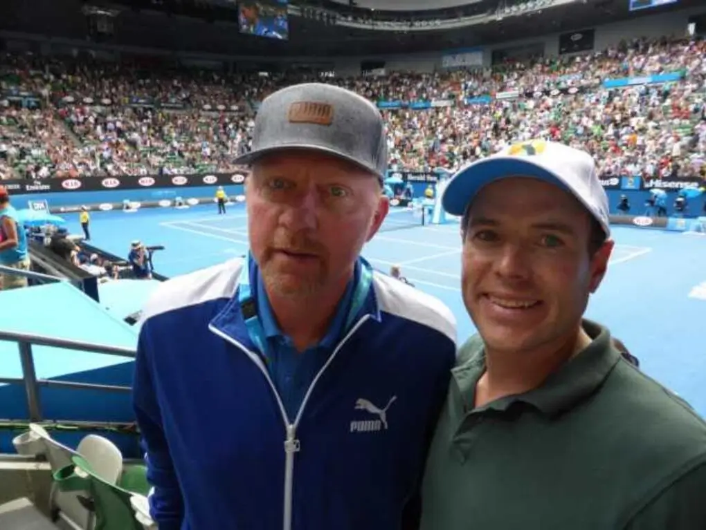 A close-up photo of tennis legend Boris Becker in a blue and white Puma tracksuit and grey cap, standing next to a smiling man in a green polo shirt at a crowded tennis stadium.