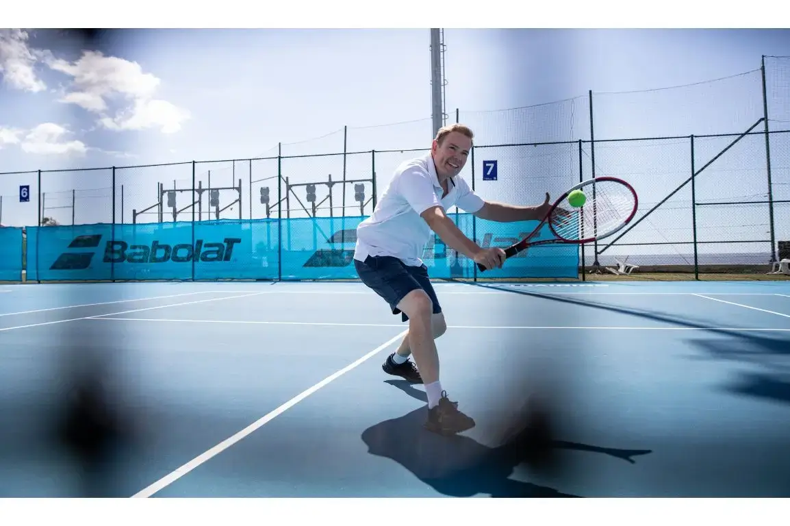 A man in a white polo shirt and dark shorts hits a tennis ball with a red racket on a bright blue hard court with Babolat windbreaks in the background.
