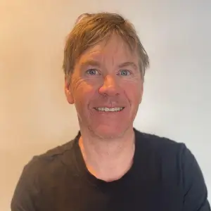 Professional portrait of a man with light brown hair wearing a black t-shirt, set against a plain neutral background.