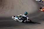 A kart racer in a white and blue suit and green helmet leaning into a sharp turn on an asphalt track.