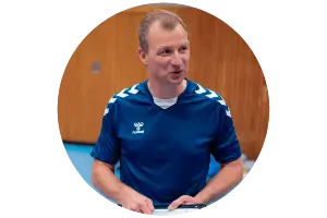 A circular-cropped headshot of a man in a blue athletic jersey with white shoulder detailing, holding a clipboard in an indoor gym.