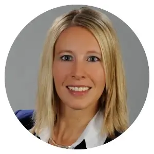 A circular professional headshot of a smiling woman with blonde hair, wearing a white collared shirt and a blue blazer against a neutral grey background.