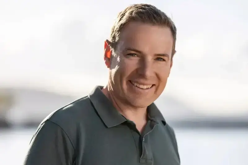 A smiling man in a dark olive green polo shirt posing for a portrait outdoors with a soft-focus background.