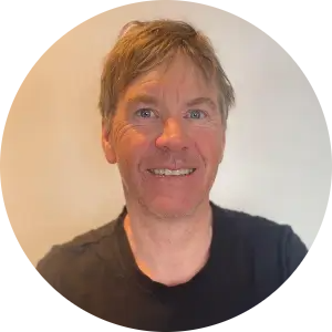 A circular-cropped headshot of a smiling man with sandy blonde hair wearing a black t-shirt against a neutral background.