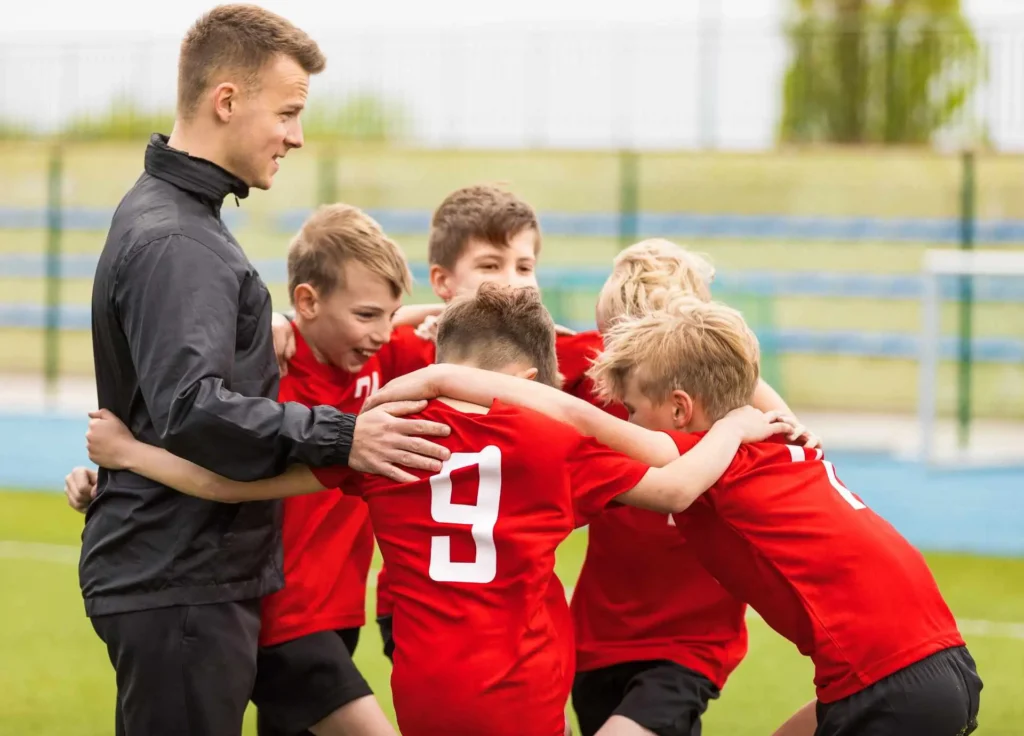 A young adult coach in a black jacket huddling with a group of youth soccer players wearing red jerseys on a grass field.