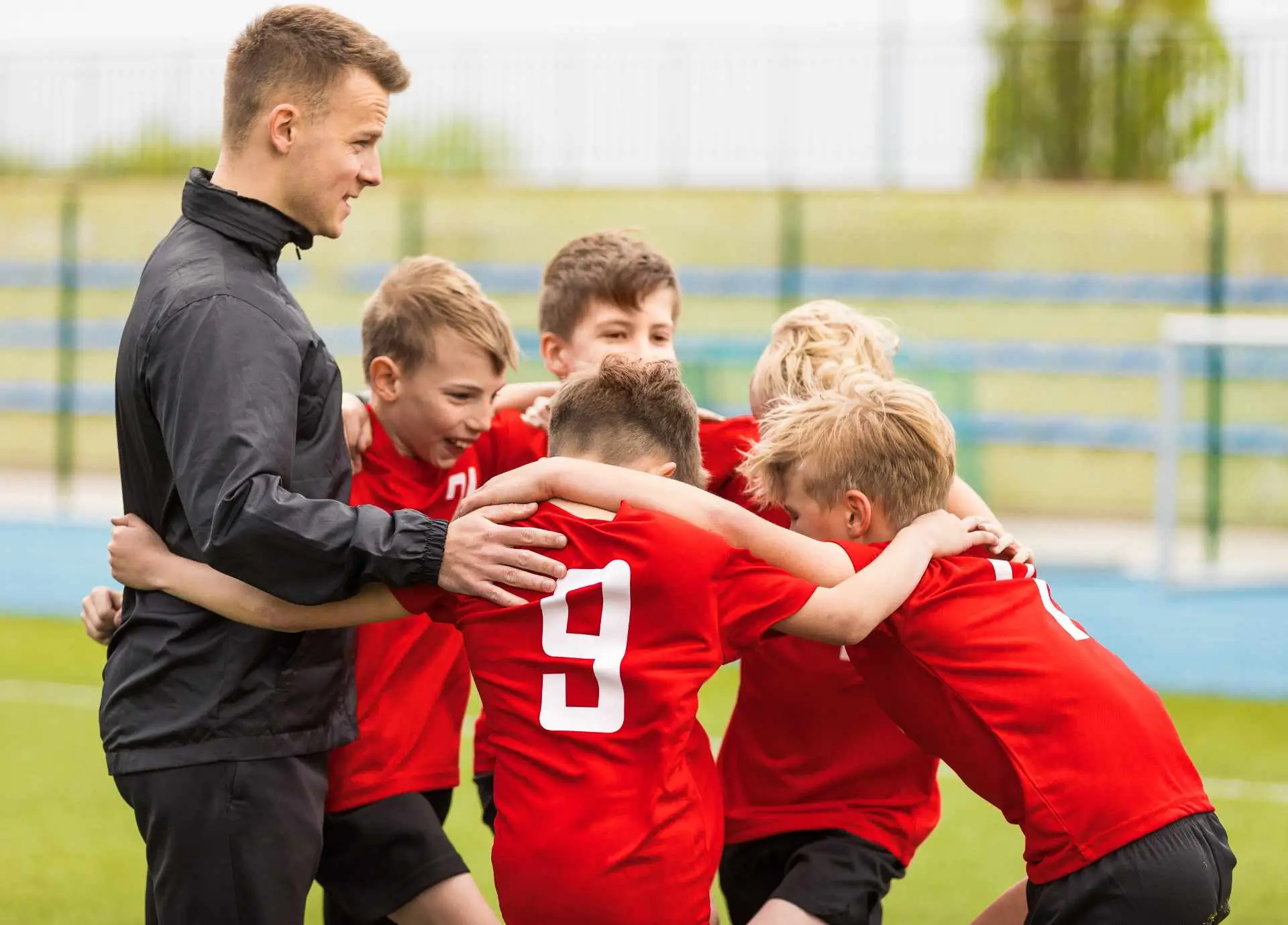 A young adult coach in a black jacket huddling with a group of youth soccer players wearing red jerseys on a grass field.