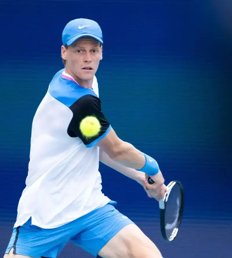 A tennis player in a blue cap and white-and-black athletic shirt prepares a two-handed backhand shot against a solid blue court background.