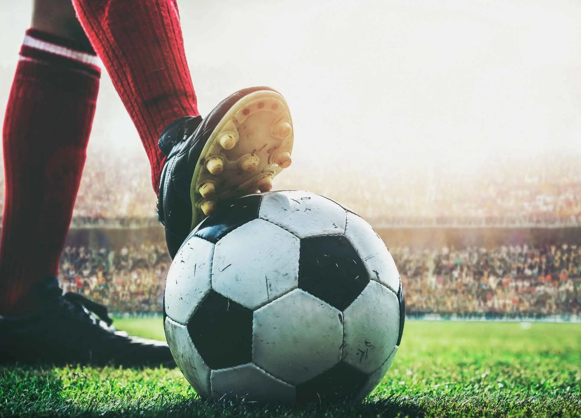 A close-up of a soccer player's cleat resting on a ball, with a blurred stadium crowd and bright field lights in the background.