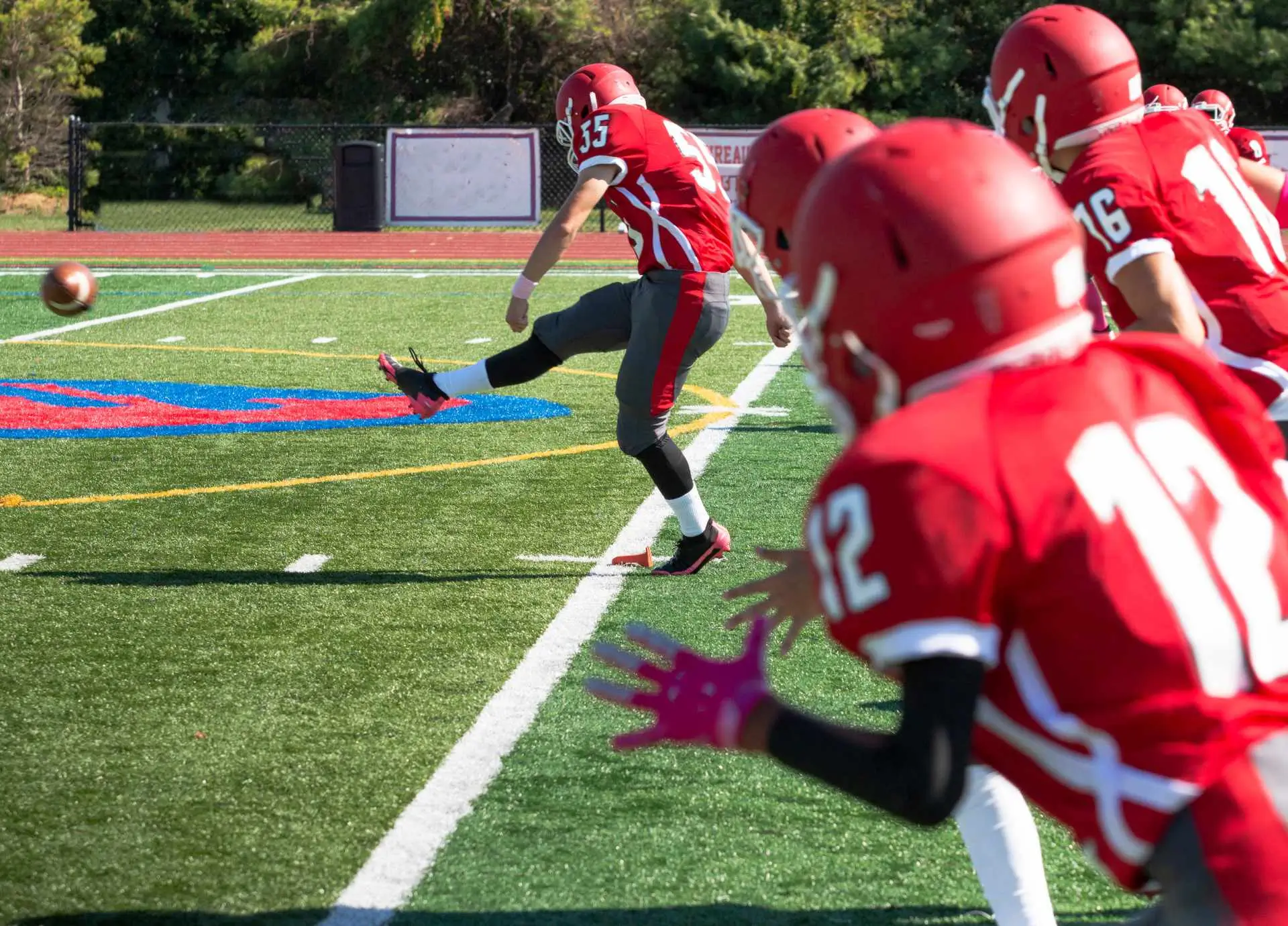 An American football player in a red jersey kicking a football on a green field during a game.