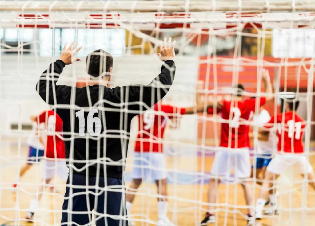 A goalkeeper wearing number 16 stands in front of a sports goal net, with a team of players in red jerseys blurred in the background.