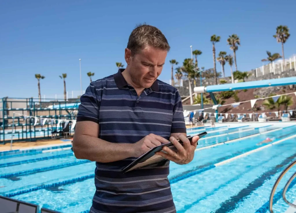 A professional swim coach in a striped polo shirt holds a clipboard while standing beside an outdoor swimming pool facility on a sunny day.