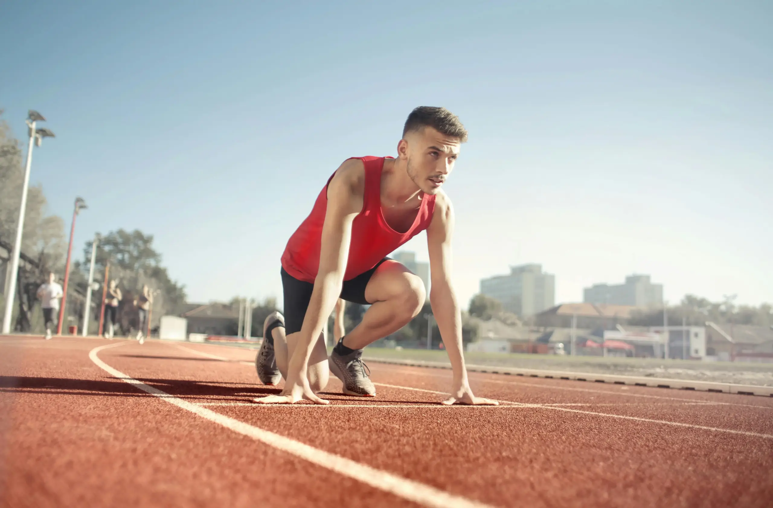 A young male runner in a red tank top in the starting position on an outdoor running track.