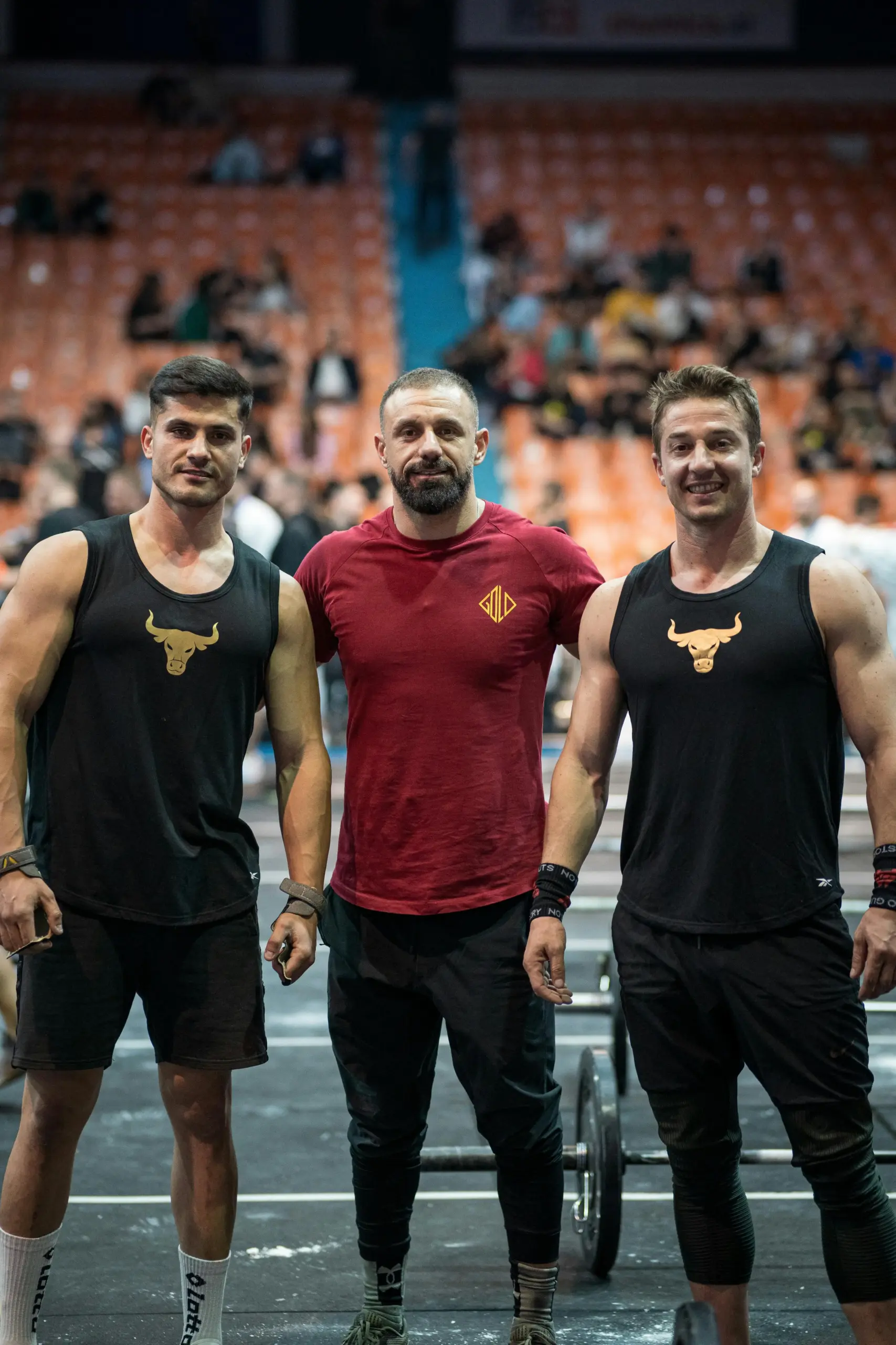 Three athletic men posing together inside a sports arena or fitness competition venue.